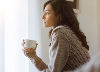 A woman holds a cup of coffee, looking out a window and thinking.