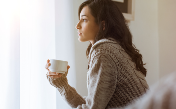 A woman holds a cup of coffee, looking out a window and thinking.