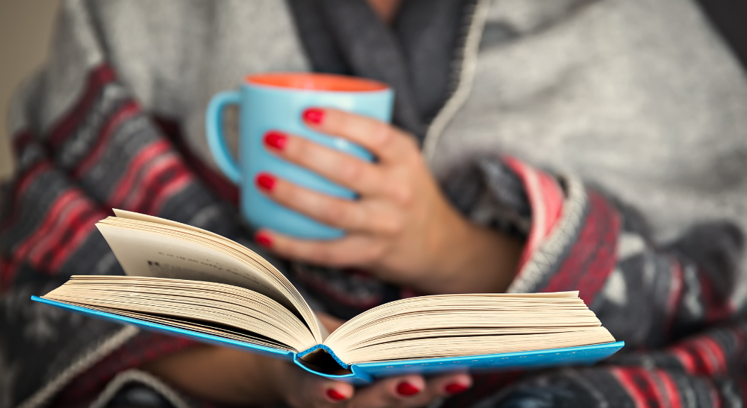 A woman holds a book and a mug of coffee or tea, while wrapped in a cozy blanket.