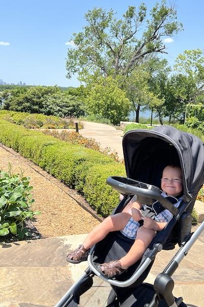 Sally Ann Rivera's toddler sits smiling in a stroller in front of the vegetable garden at the Dallas Arboretum.