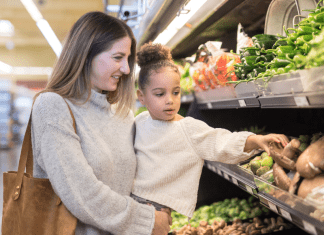 A mom holds her daughter as she reaches for a mushroom in the produce section of a grocery store.