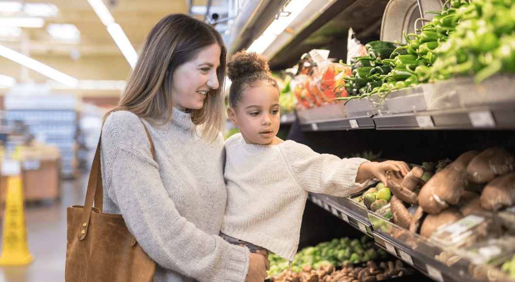 A mom holds her daughter as she reaches for a mushroom in the produce section of a grocery store.