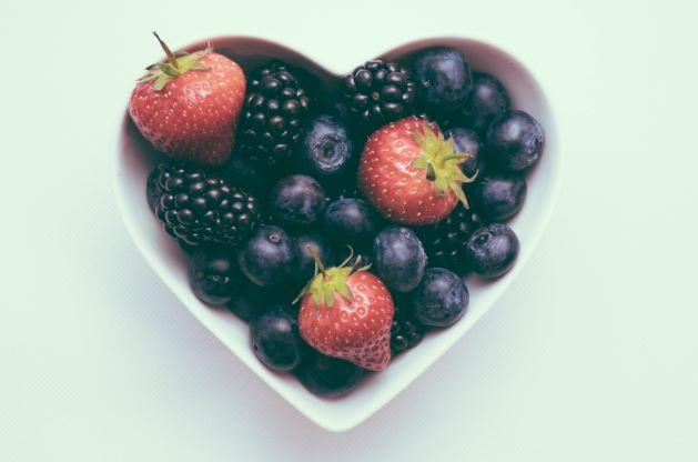 A white, heart shaped ceramic bowl holds a mix of strawberries, blackberries, and blueberries.