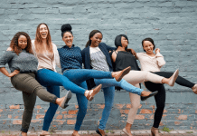 Six women of all types of skin colors locking arms and kicking one leg in the air, laughing