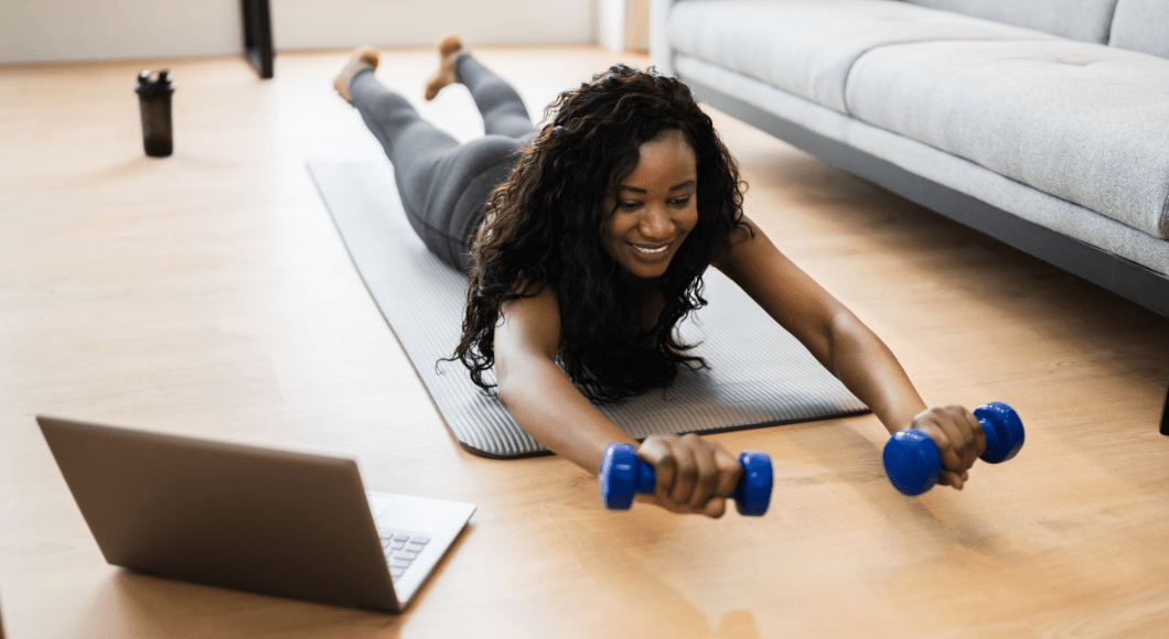 A woman workouts on on a yoga mat with two weights in her hands and watching her laptop.