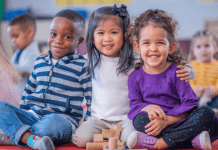 Three preschoolers sit on the ground with the middle child embracing them.