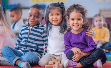 Three preschoolers sit on the ground with the middle child embracing them.