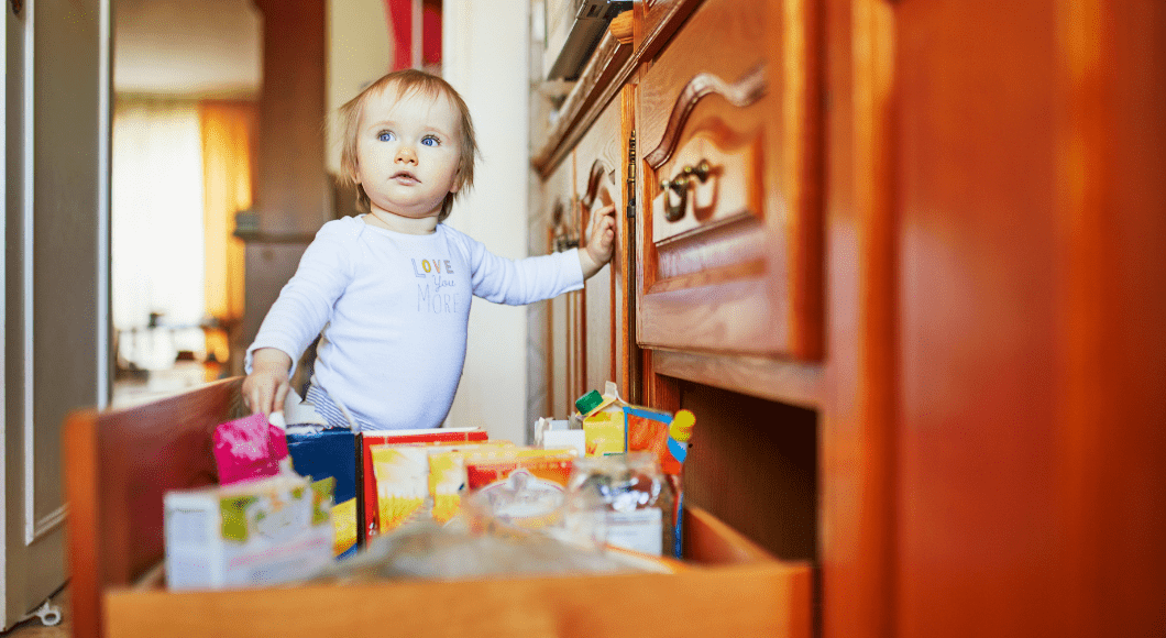 A baby stands in a kitchen with a drawer open.