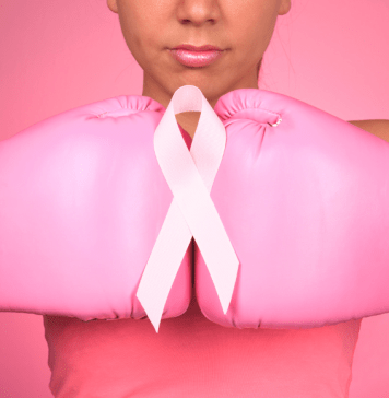 A woman wears two pink boxing gloves with a breast cancer ribbon on top.