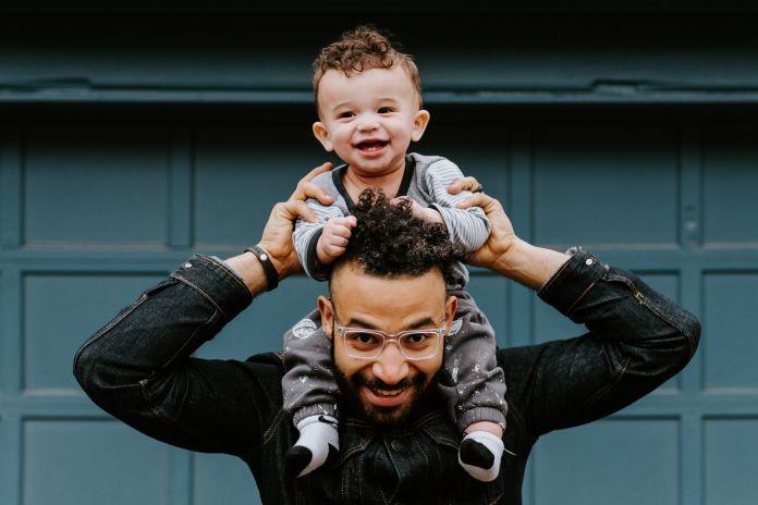 A dad wearing glasses holds his toddler son on his shoulder while they both smile and laugh.