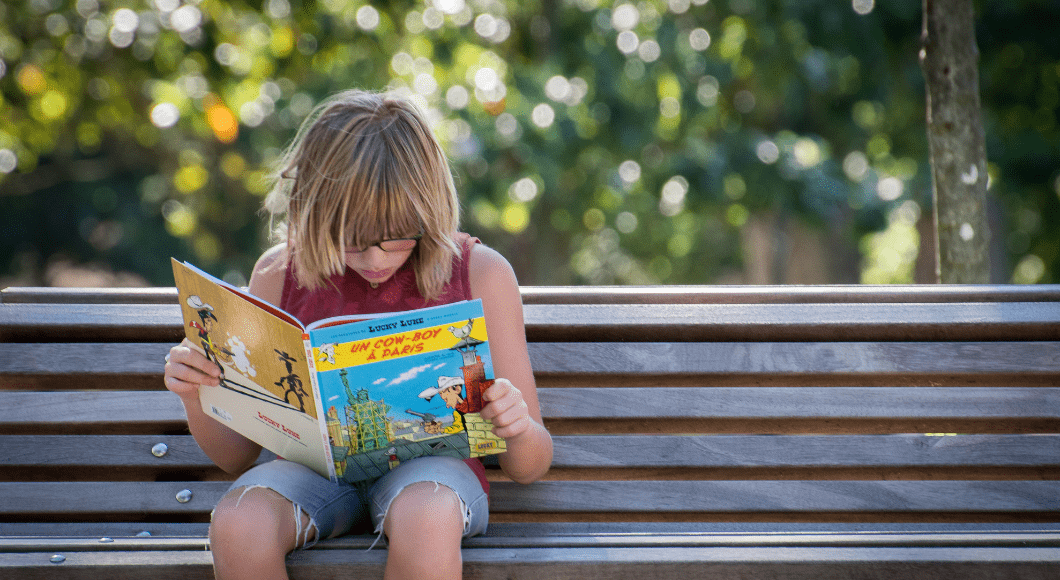 A young girl reads a book on a bench outside.