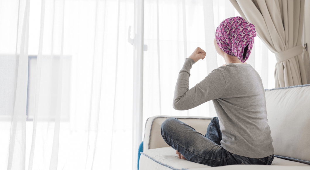 Breast cancer patient sits on a couch at her house looking out a window and flexing her arm.