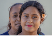 A Latina mom stands behind her daughter, who has a flower in her hair.