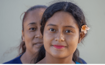 A Latina mom stands behind her daughter, who has a flower in her hair.