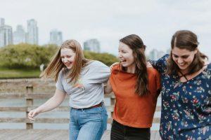Three ladies laugh while walking arm in arm across a bridge with a skyline behind them.