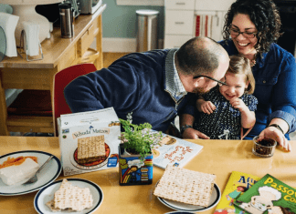 A mom, dad, and young daughter celebreate passover with books and matzah on a table